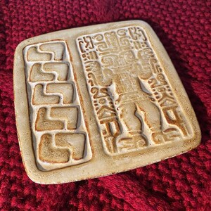 May include: A beige ceramic tile with a raised design of a Mayan figure and glyphs. The figure is wearing a headdress and a loincloth. The glyphs are in a stylized script.