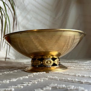 May include: A gold-toned metal bowl with a decorative floral design around the base. The bowl has a hammered texture and sits on a pedestal.