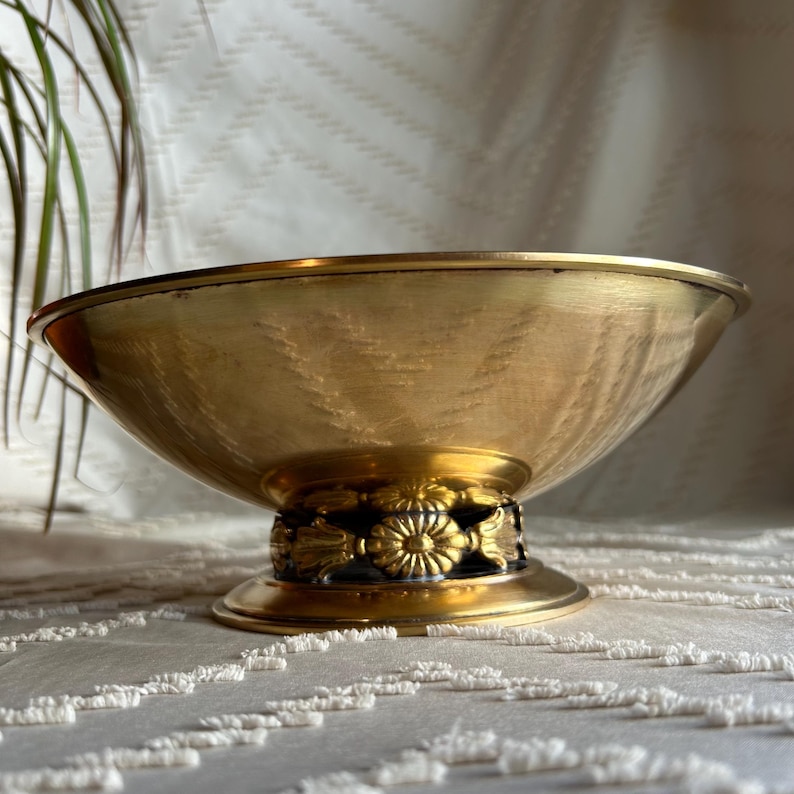May include: A gold-toned metal bowl with a decorative floral design around the base. The bowl has a hammered texture and sits on a pedestal.