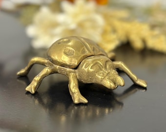 May include: A brass beetle figurine with a hinged top, showcasing intricate details. The beetle's body is segmented, and it has six legs. The figurine is a decorative item, likely for display or as a small trinket box.
