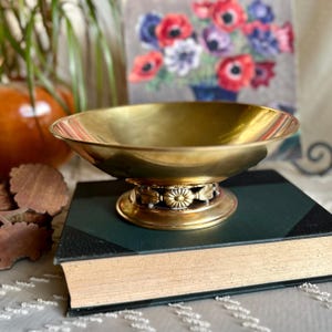 May include: A gold-toned metal bowl with a decorative floral design around the base. The bowl is sitting on a dark green book.
