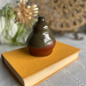 May include: A small, pear-shaped ceramic bud vase with a glossy finish. The upper portion of the vase features a speckled dark olive green glaze, while the lower base is a solid, smooth terracotta red. A single dried flower is placed inside.