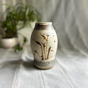 May include: A ceramic vase with a speckled off-white glaze featuring a dark brown painted botanical design. The vase has a tapered neck and a wider body, with a dark grey speckled band at the base and a dark brown speckled rim at the top opening.
