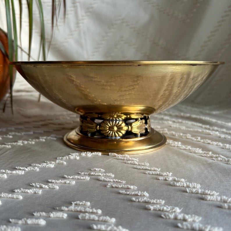 May include: A gold-toned metal bowl with a decorative base. The bowl has a smooth, shiny surface and a slightly flared rim. The base features a ring of ornate floral designs.
