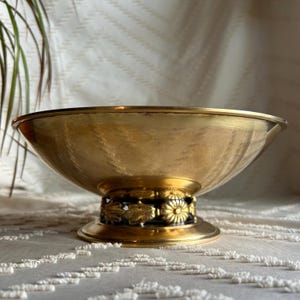 May include: A gold-toned metal bowl with a decorative base featuring black and gold floral accents. The bowl is round and has a smooth, polished surface.