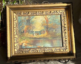 May include: A framed oil painting depicting a woodland scene with a river, trees, and figures in the distance. The frame is gold with an ornate design.