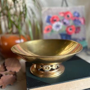 May include: A gold-toned brass bowl with a decorative base sits on top of a closed book. The bowl has a round shape and a smooth, reflective surface.