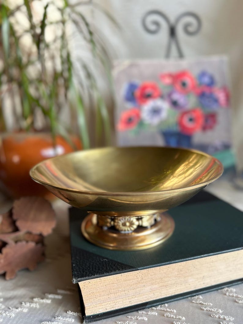 May include: A gold-toned brass bowl with a decorative base sits on top of a closed book. The bowl has a round shape and a smooth, reflective surface.