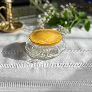 May include: A vintage glass and amber powder compact with a decorative design. The compact is closed and sitting on a white surface.