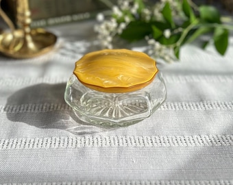 May include: A vintage glass and amber powder compact with a decorative design. The compact is closed and sitting on a white surface.