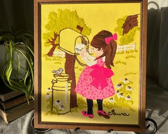 May include: A framed embroidered picture of a girl in a pink dress with white polka dots, standing by a yellow mailbox and holding a letter. The background is a yellow landscape with a blue bird and a white picket fence. The picture is signed "Laura".