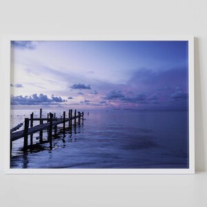 May include: A framed print depicting a tranquil seascape at twilight. A wooden pier stretches into the still water, mirroring the soft colours of the sky. The sky transitions from purple to blue, with scattered clouds. The print is in a white frame.