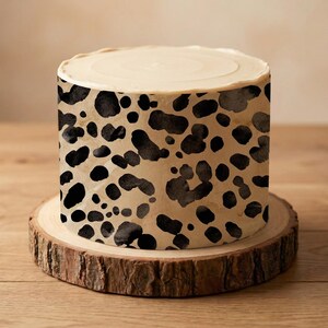 May include: A round cake with pale frosting and a brown and black spotted pattern around the sides. The cake sits on a round, natural wood slice. The background is light brown.