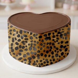 May include: A heart-shaped cake with a chocolate topping. The sides of the cake feature a brown and black leopard print design. The cake is presented on a white cake stand.
