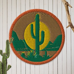 Puede incluir: Un parche redondo bordado con un fondo marrón, un cactus verde, un sol amarillo y montañas verdes. El parche tiene un borde naranja.