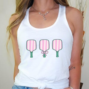 Pode incluir: Camisola branca de al&ccedil;as com um gr&aacute;fico de tr&ecirc;s raquetes de paddleball listradas de rosa e verde. A raquete do meio tem um la&ccedil;o rosa.