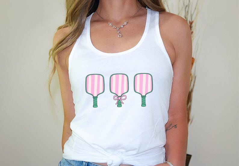 Pode incluir: Camisola branca de al&ccedil;as com um gr&aacute;fico de tr&ecirc;s raquetes de paddleball listradas de rosa e verde. A raquete do meio tem um la&ccedil;o rosa.