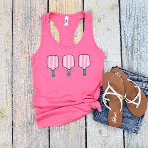 Pode incluir: Camisola de al&ccedil;as rosa com um gr&aacute;fico de tr&ecirc;s raquetes de paddlebol rosa e branco com al&ccedil;as verdes e um la&ccedil;o rosa na raquete do meio.