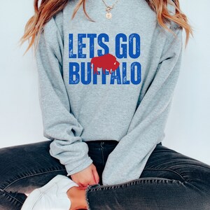 May include: A gray sweatshirt with the text "LETS GO BUFFALO" in blue with a red buffalo silhouette.