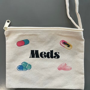 Puede incluir: Una bolsa de lona blanca con cremallera y asa negra. La bolsa tiene la palabra "Meds" impresa en negro y varias ilustraciones de p&iacute;ldoras de colores.