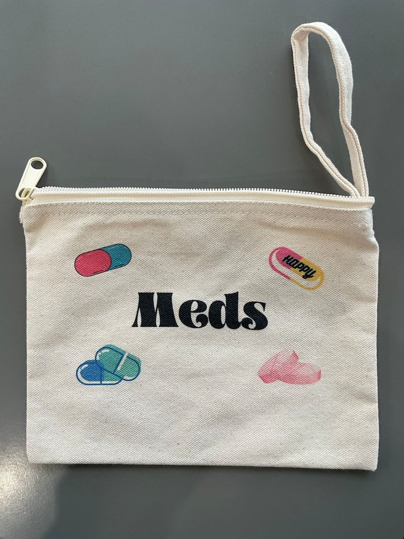 Puede incluir: Una bolsa de lona blanca con cremallera y asa negra. La bolsa tiene la palabra "Meds" impresa en negro y varias ilustraciones de p&iacute;ldoras de colores.