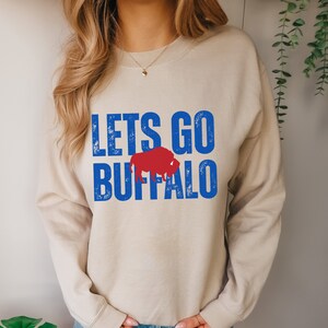 May include: A beige sweatshirt with the text "LETS GO BUFFALO" in blue and red. The text is in a distressed font and a red buffalo silhouette is between the words "GO" and "BUFFALO".