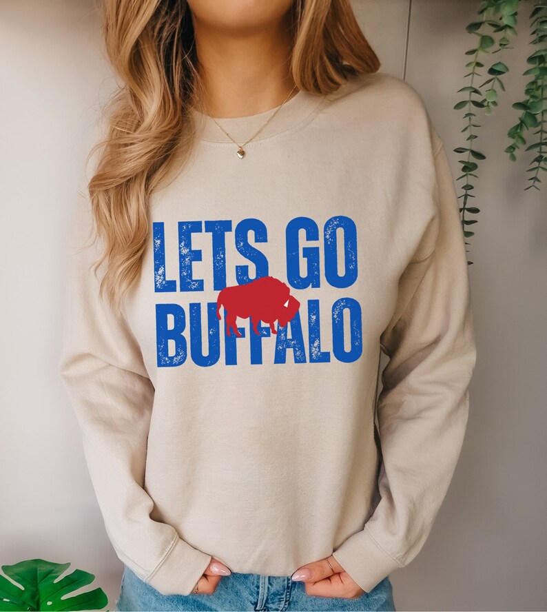 May include: A beige sweatshirt with the text "LETS GO BUFFALO" in blue and red. The text is in a distressed font and a red buffalo silhouette is between the words "GO" and "BUFFALO".