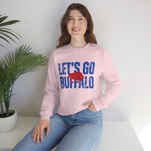 May include: A pink sweatshirt with the text "Let's Go Buffalo" in blue with a red buffalo silhouette.