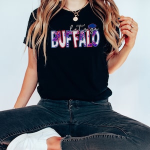 May include: A black t-shirt with the text "let's go BUFFALO" in red, white, and blue. The letters are outlined in white and have a distressed look.