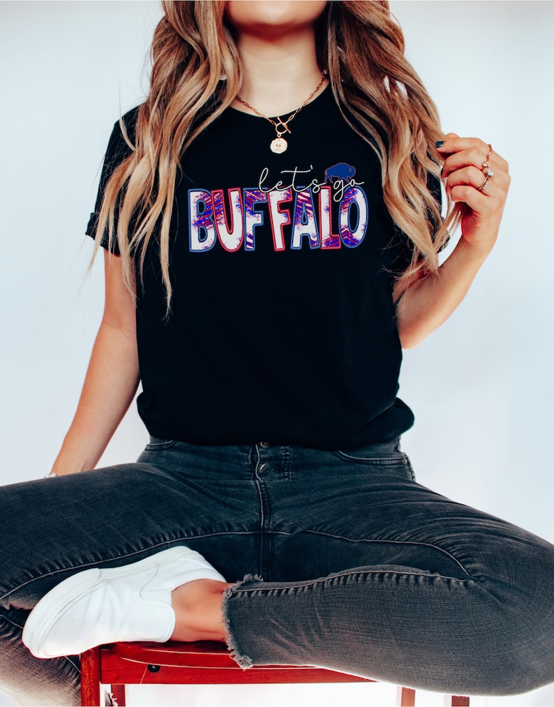May include: A black t-shirt with the text "let's go BUFFALO" in red, white, and blue. The letters are outlined in white and have a distressed look.