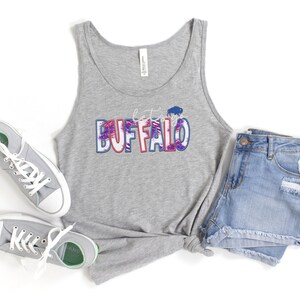 May include: A gray tank top with the text "Let's go Buffalo" in red, white, and blue with a buffalo silhouette. The tank top is paired with denim shorts and gray sneakers.