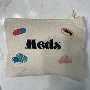 Puede incluir: Una bolsa de lona blanca con cremallera con la palabra "Meds" impresa en negro. La bolsa tiene dise&ntilde;os coloridos en forma de p&iacute;ldora con la palabra "Happy" impresa en una de las p&iacute;ldoras.