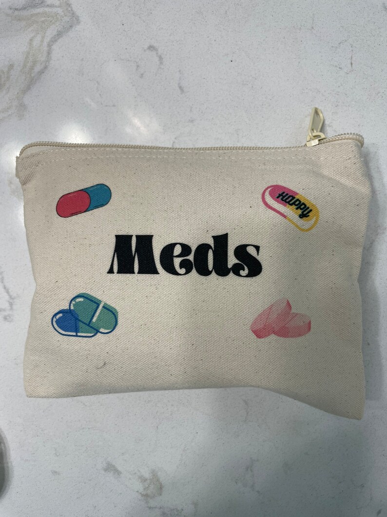 Puede incluir: Una bolsa de lona blanca con cremallera con la palabra "Meds" impresa en negro. La bolsa tiene dise&ntilde;os coloridos en forma de p&iacute;ldora con la palabra "Happy" impresa en una de las p&iacute;ldoras.