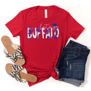 May include: Red t-shirt with the text "let's go BUFFALO" in blue and pink with a white buffalo silhouette inside the "O". The shirt is styled with denim shorts and striped sandals.