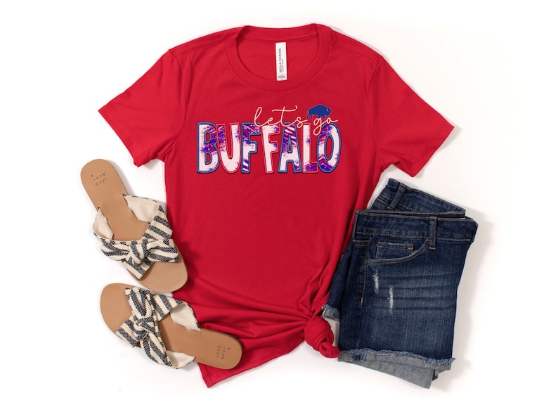 May include: Red t-shirt with the text "let's go BUFFALO" in blue and pink with a white buffalo silhouette inside the "O". The shirt is styled with denim shorts and striped sandals.