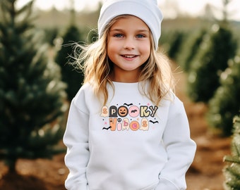 Puede incluir: Una persona joven con una sudadera blanca con las palabras "Spooky Vibes" en letras coloridas y gráficos temáticos de Halloween. La persona también lleva un gorro blanco. El fondo es una escena borrosa al aire libre con árboles verdes.