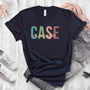 Könnte beinhalten: Marineblaues T-Shirt mit dem Text "CASE manager" in bunten Buchstaben. Die Buchstaben sind in Grün-, Blau-, Rosa- und Orangetönen gehalten.