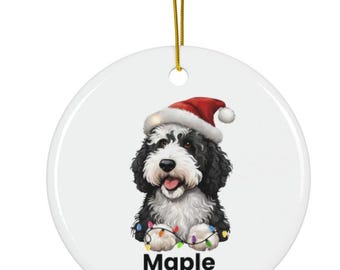 Könnte beinhalten: Runder, weißer Weihnachtsschmuck mit einem schwarz-weißen Hund, der einen Weihnachtsmannhut trägt. Der Hund ist von bunten Weihnachtslichtern umgeben. Der Name "Maple" und das Jahr "2025" sind unten in Schwarz gedruckt.