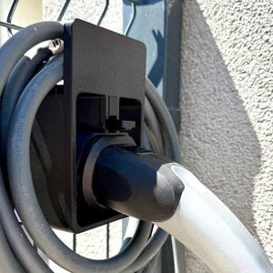 Può includere: Stazione di ricarica nera per veicoli elettrici, montata su un muro strutturato. La stazione include una porta di ricarica nera e un cavo di ricarica grigio avvolto attorno. Il cavo è collegato a un'impugnatura di ricarica grigia.