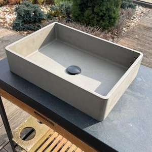 Può includere: Un lavandino rettangolare in cemento grigio chiaro con uno scarico nero. Il lavandino è appoggiato su una superficie grigio scuro, con un ripiano in legno a doghe sottostante. L'ambiente esterno comprende vegetazione e una piscina.