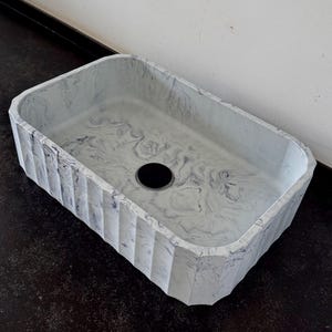 Può includere: Lavandino rettangolare da bagno con motivo marmorizzato grigio e bianco. Il lavandino ha un foro di scarico nero e un esterno strutturato e scanalato. Il lavandino è realizzato in un materiale solido ed è progettato per l'uso in bagno.