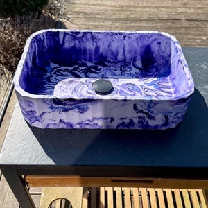 May include: A rectangular bathroom sink with a marbled purple and white pattern. The sink has a dark grey drain cover and is set on a dark grey table. The sink's design features a swirling, artistic aesthetic.