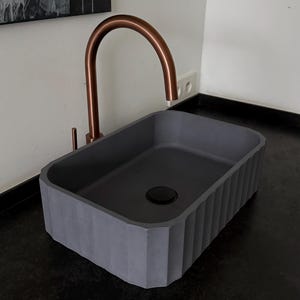 Peut inclure: Un lavabo rectangulaire en béton gris avec un robinet de couleur cuivre. L'évier a un design extérieur nervuré et un drain foncé. Le robinet a un bec courbé. L'évier est sur un comptoir noir.