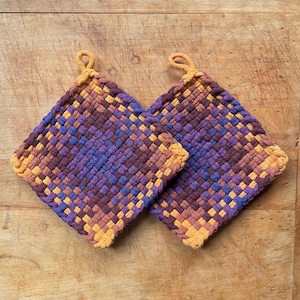 May include: Two square woven potholders in shades of purple, brown, and orange. Each potholder has a loop for hanging and a woven design. The potholders are handmade and suitable for kitchen use.