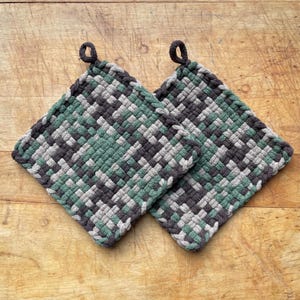 May include: Two square woven potholders in shades of green, grey, and black. Each potholder has a loop for hanging. The potholders are made of a thick, textured material and are placed on a wooden surface.