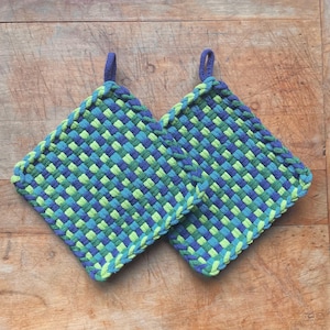 May include: Two square woven potholders in shades of blue and green. Each potholder has a loop for hanging and a braided edge. The woven pattern creates a textured appearance, perfect for kitchen use.