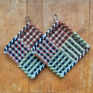 May include: Two square woven pot holders with a loop for hanging. The pot holders feature a woven pattern in shades of brown, blue, green, and red. The edges are trimmed with a braided rope in a neutral color. They are sitting on a wooden surface.