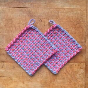 May include: Two square woven potholders in pink, red, and blue-grey hues. Each potholder has a loop for hanging and a braided edge. The potholders are set on a wooden surface.