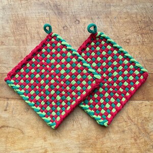 May include: Two woven potholders in a red and green checkerboard pattern. Each potholder has a loop for hanging and a braided green border. The potholders are square and appear to be handmade.