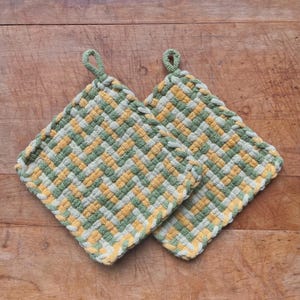 May include: Two square woven potholders in a basket weave pattern. The potholders feature a repeating pattern of green, yellow, and cream-colored threads. Each potholder has a small green loop for hanging. The potholders are on a wooden surface.